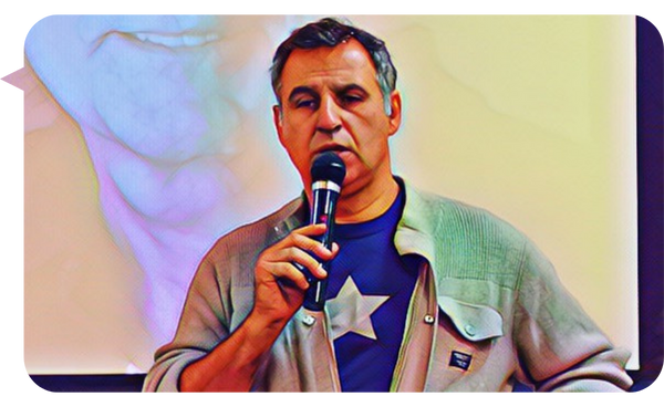 Marcelo Borlando speaking into a microphone during a presentation, wearing a star-themed shirt and a light jacket, with his image projected in the background.